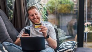 Guy relaxing on couch with laptop in his lap and credit card and mobile phone in either hand.