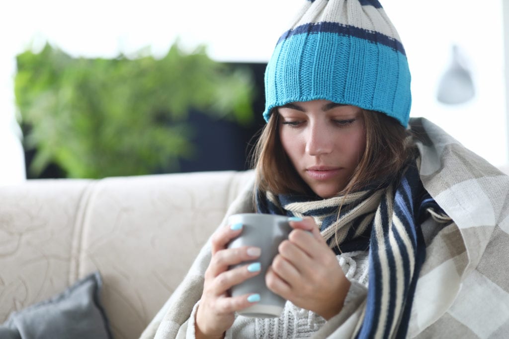 Close-up of sad ill woman drinking tea for keep warmly. Female wrapping in warm blanket on sick leaves at home. Seasonal sickness and treatment concept