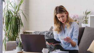Woman sitting on couch holding calculator and bill with coffee table in front of her holding a laptop and notebook.